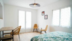 Une chambre à coucher comprenant un lit, un bureau et deux chaises. Il y a deux fenêtres équipées de stores et un luminaire au plafond.