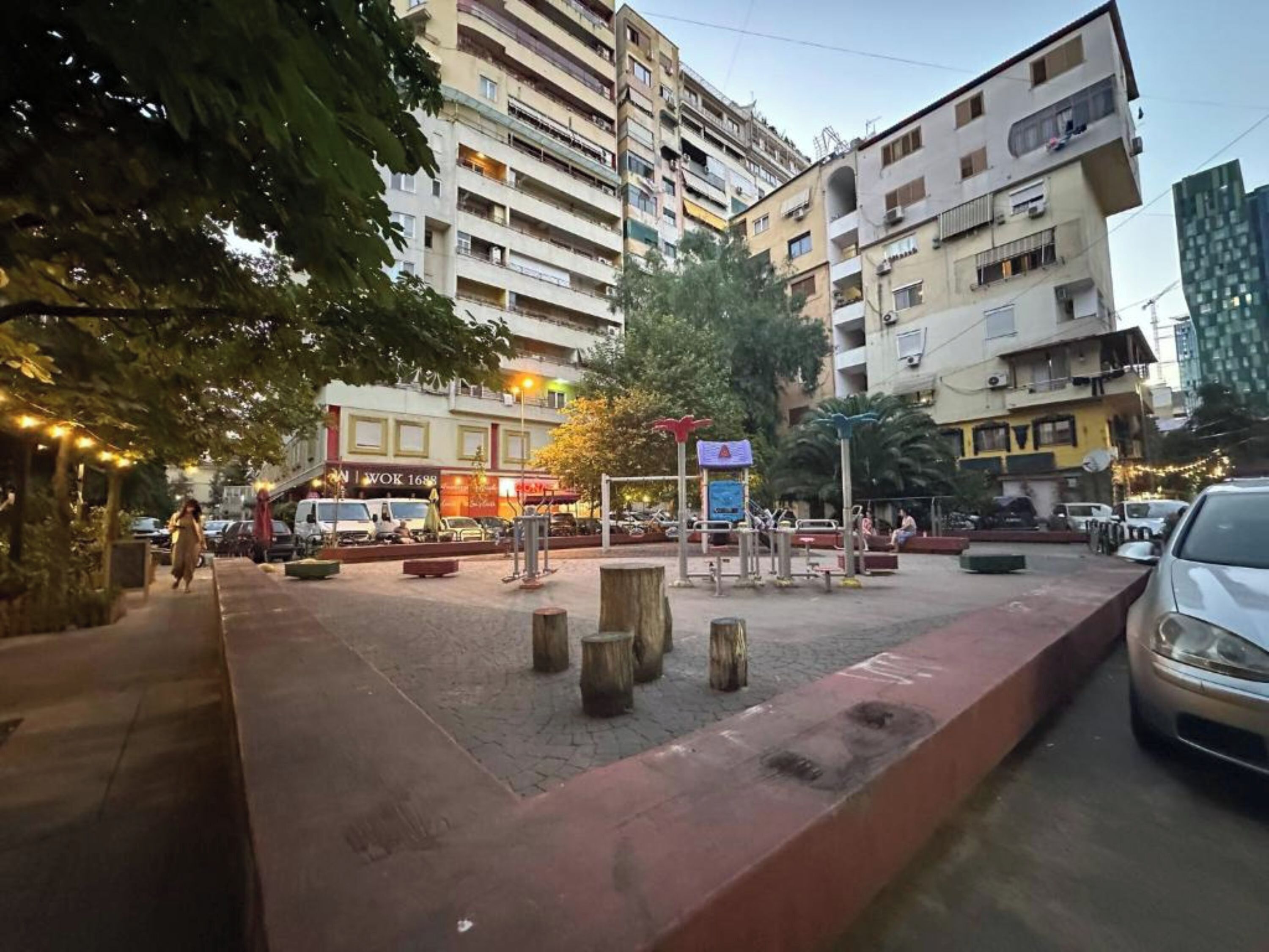 Μια πλατεία στην πόλη με παγκάκια, μια παιδική χαρά και ένα κτίριο με πολλούς ορόφους.