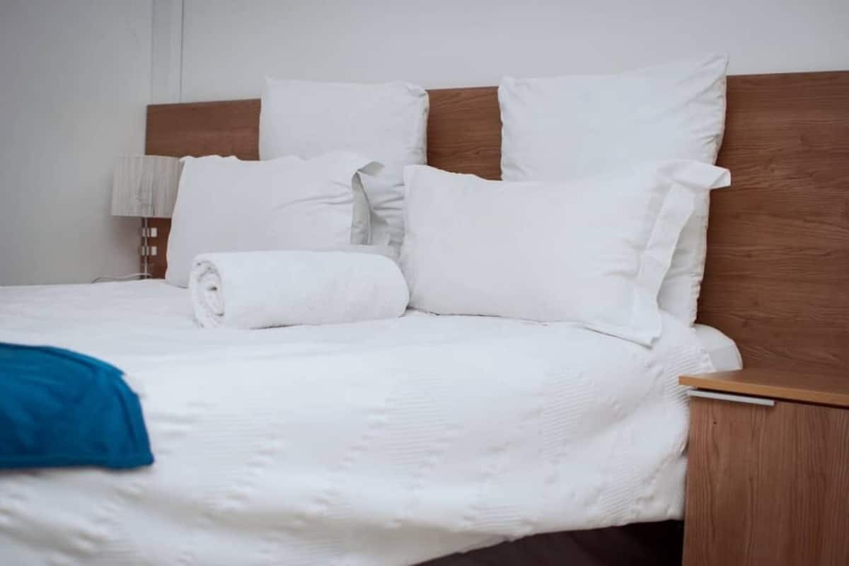 Una cama bien hecha con ropa de cama y almohadas blancas, un cabecero de madera y una mesita de noche con una lámpara.
