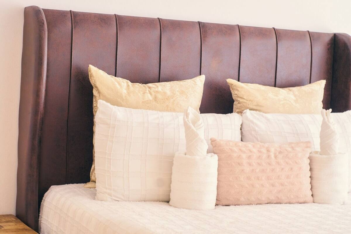 Una cama con cabecera oscura, varios almohadones y una colcha blanca.