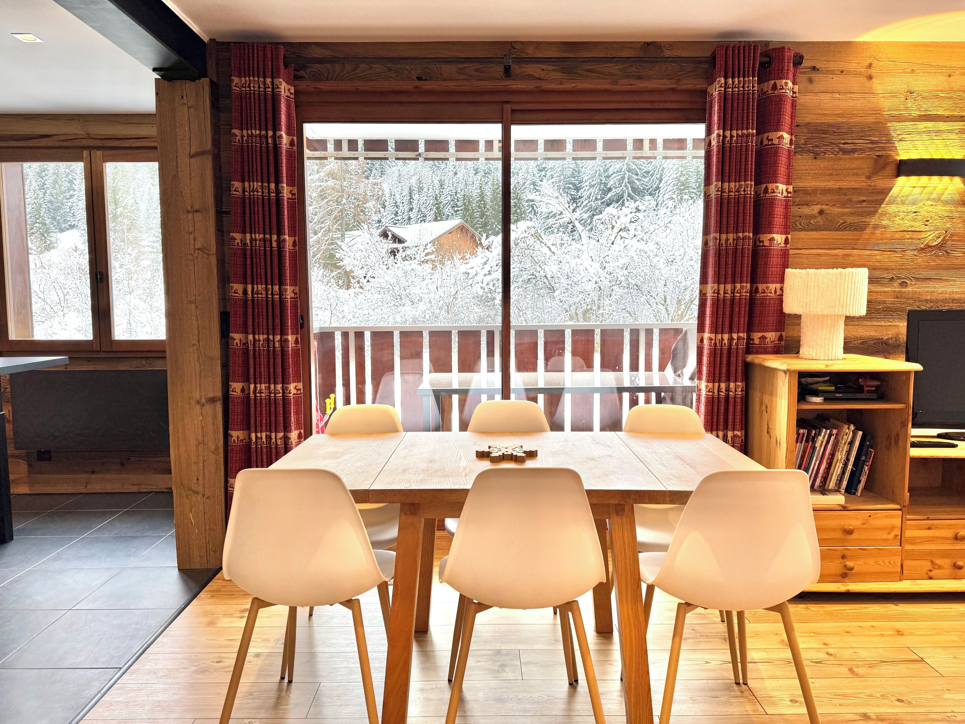 A dining area with a wooden table and four chairs, a wooden bookshelf, and a view of snow-covered trees through large windows.