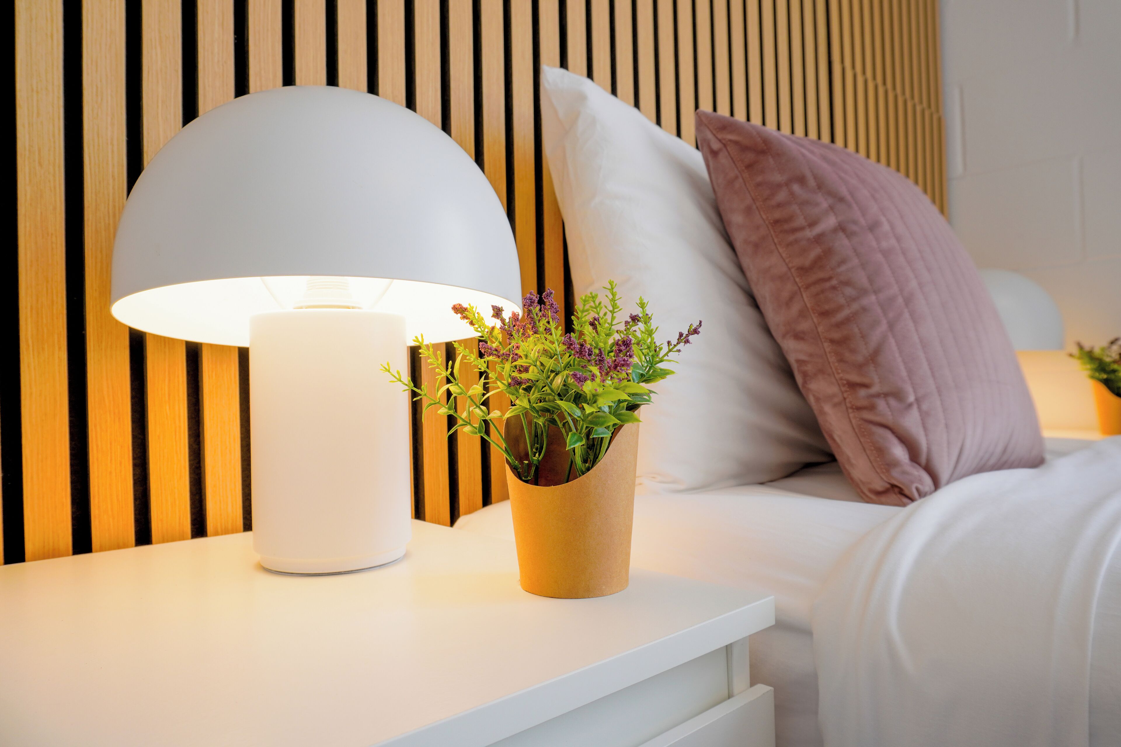 A neatly arranged bedroom with a bedside lamp, a potted plant, and a bed with pillows.