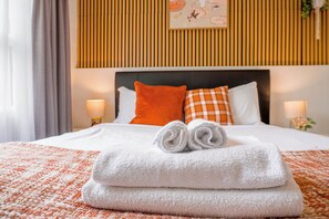 Ein ordentlich bezogenes Bett mit weißer und orangefarbener Bettwäsche, einem Kopfteil und einer Wand mit vertikalen Holzlatten.