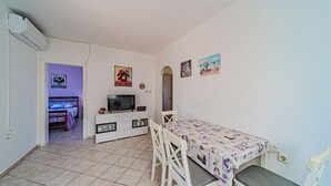 Una habitación pequeña y luminosa con una cama, un televisor, una mesa de comedor y un armario blanco.