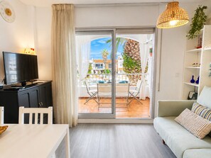 A modern living room with a television, a sofa, a dining area with a table and chairs, and a balcony with a view of buildings and palm trees. - 0146-PATTAYA Apartment with canal view (Empuriabrava)