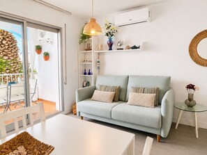 A living room with a light blue sofa, a glass coffee table, and a white dining table. There is a balcony with plants and a wicker basket. - 0146-PATTAYA Apartment with canal view (Empuriabrava)