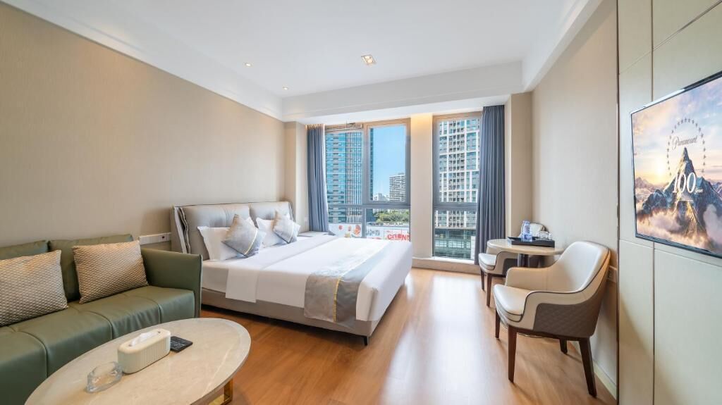 A modern hotel room with a large bed, a sofa, a small table, and a chair. There is a large window with a view of buildings outside.