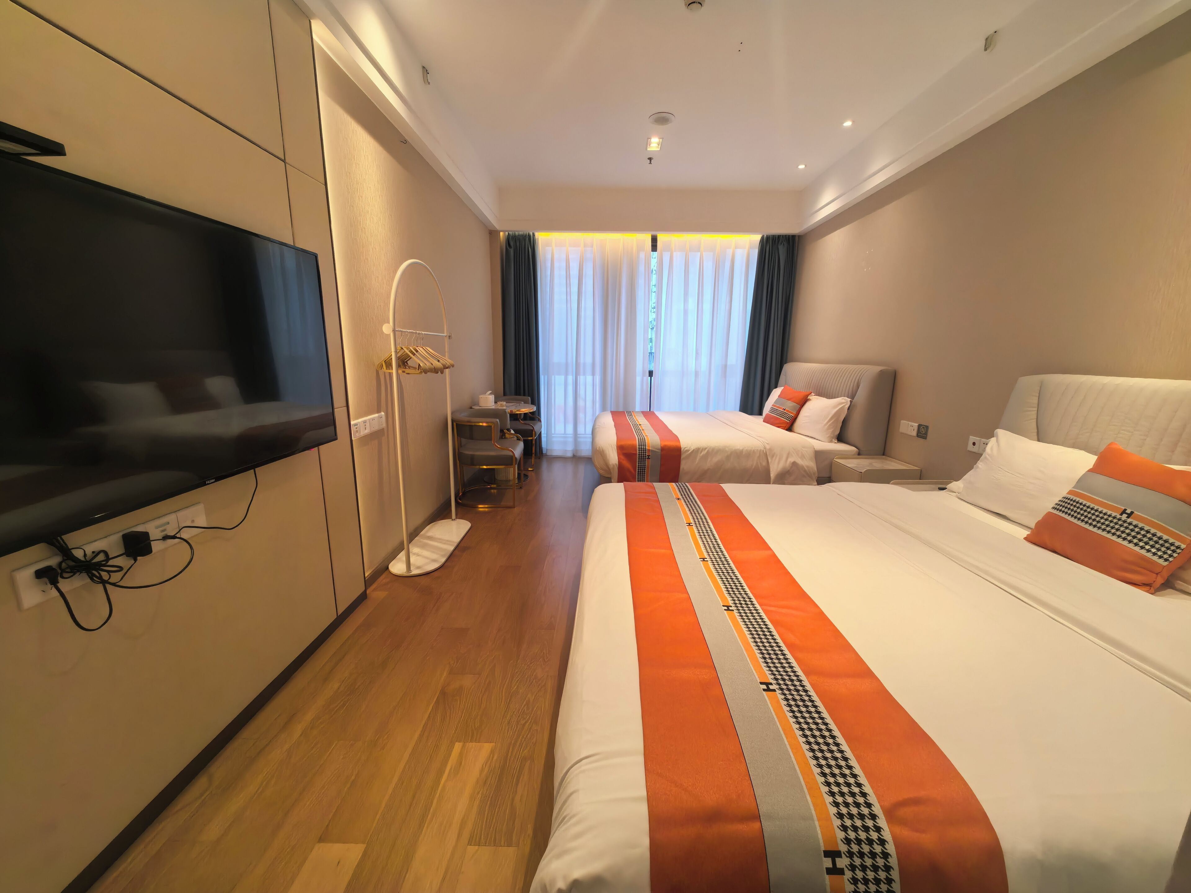 A hotel room with a flat-screen TV, two beds with orange and white bedding, a desk with chairs, and a large window with curtains.