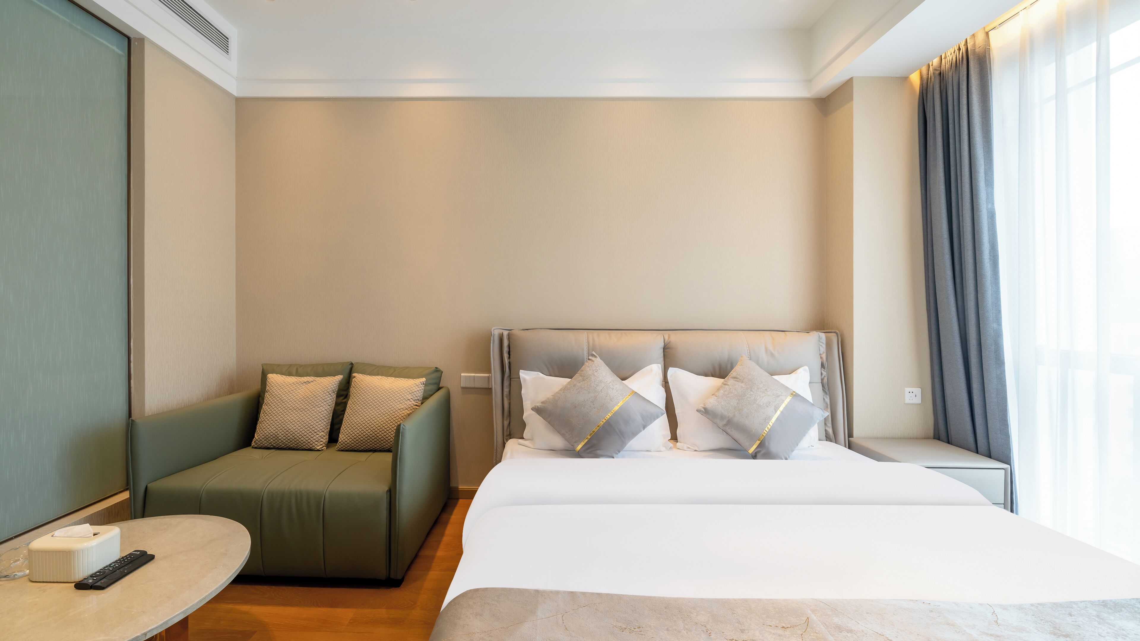 Una habitación de hotel con una cama, un sillón verde, una mesita con un control remoto y una ventana con cortinas.