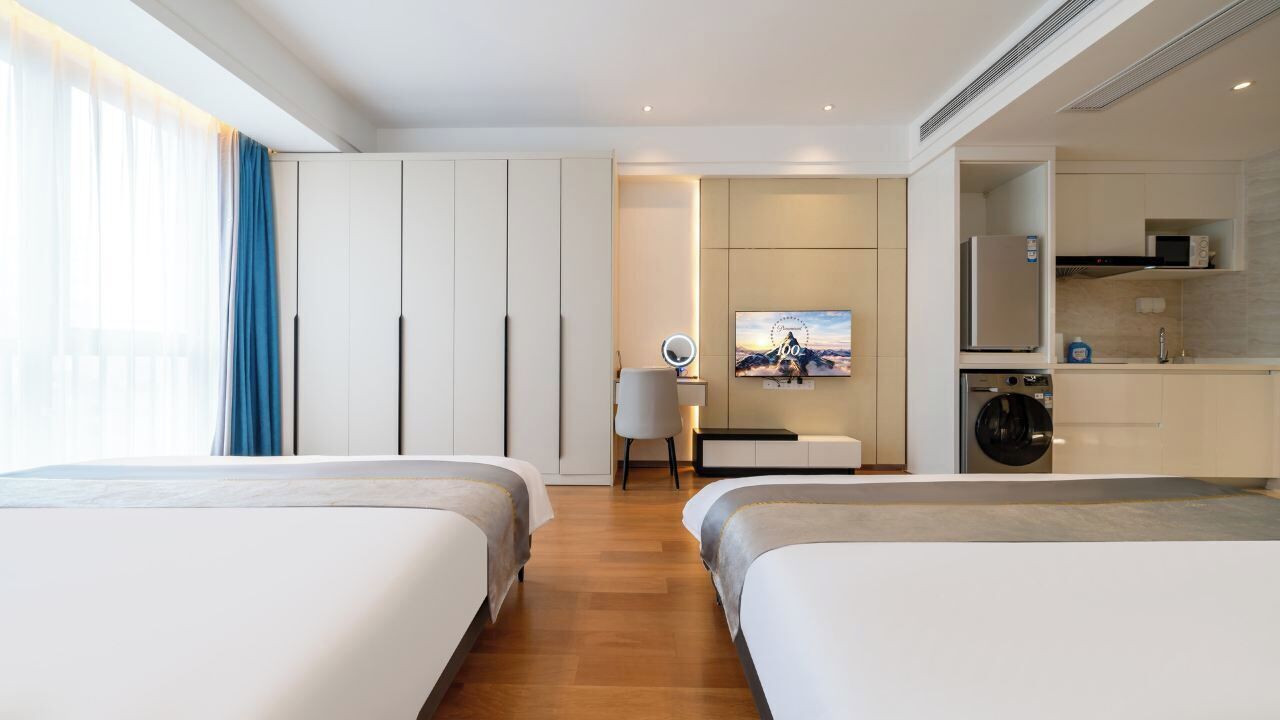A modern hotel room with two beds, a TV, a desk, and a kitchenette.