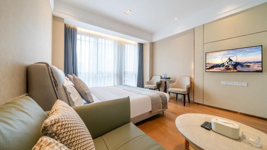 A modern hotel room with a large bed, a sofa, a small table, and a TV on the wall.