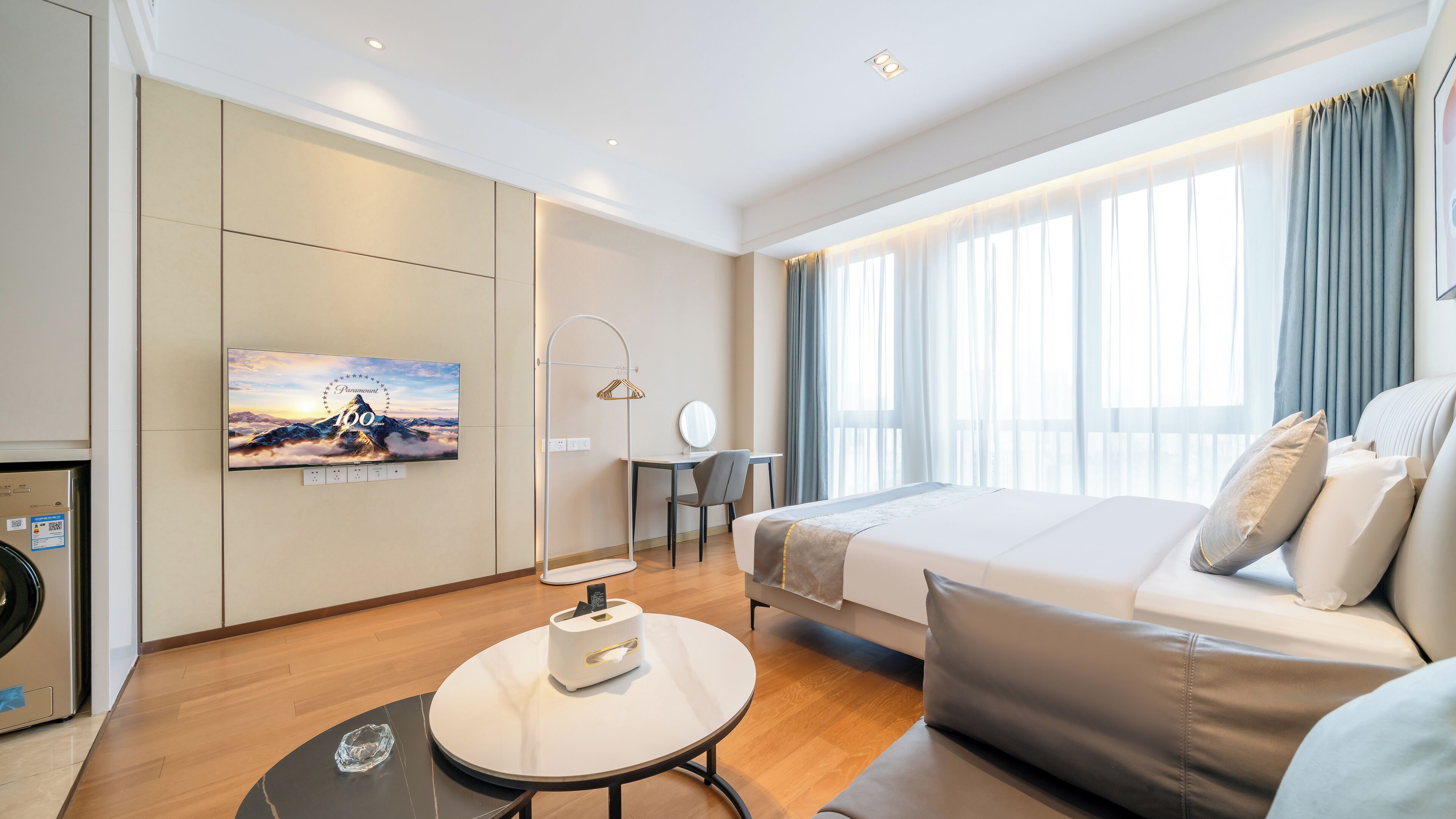 Ein modernes Hotelzimmer mit einem Bett, einem Schreibtisch, einem Stuhl, einem an der Wand befestigten Fernseher und einem Fenster mit Vorhängen.