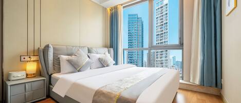 A modern bedroom with a large bed, a bedside table with a lamp, and a view of high-rise buildings through the window.