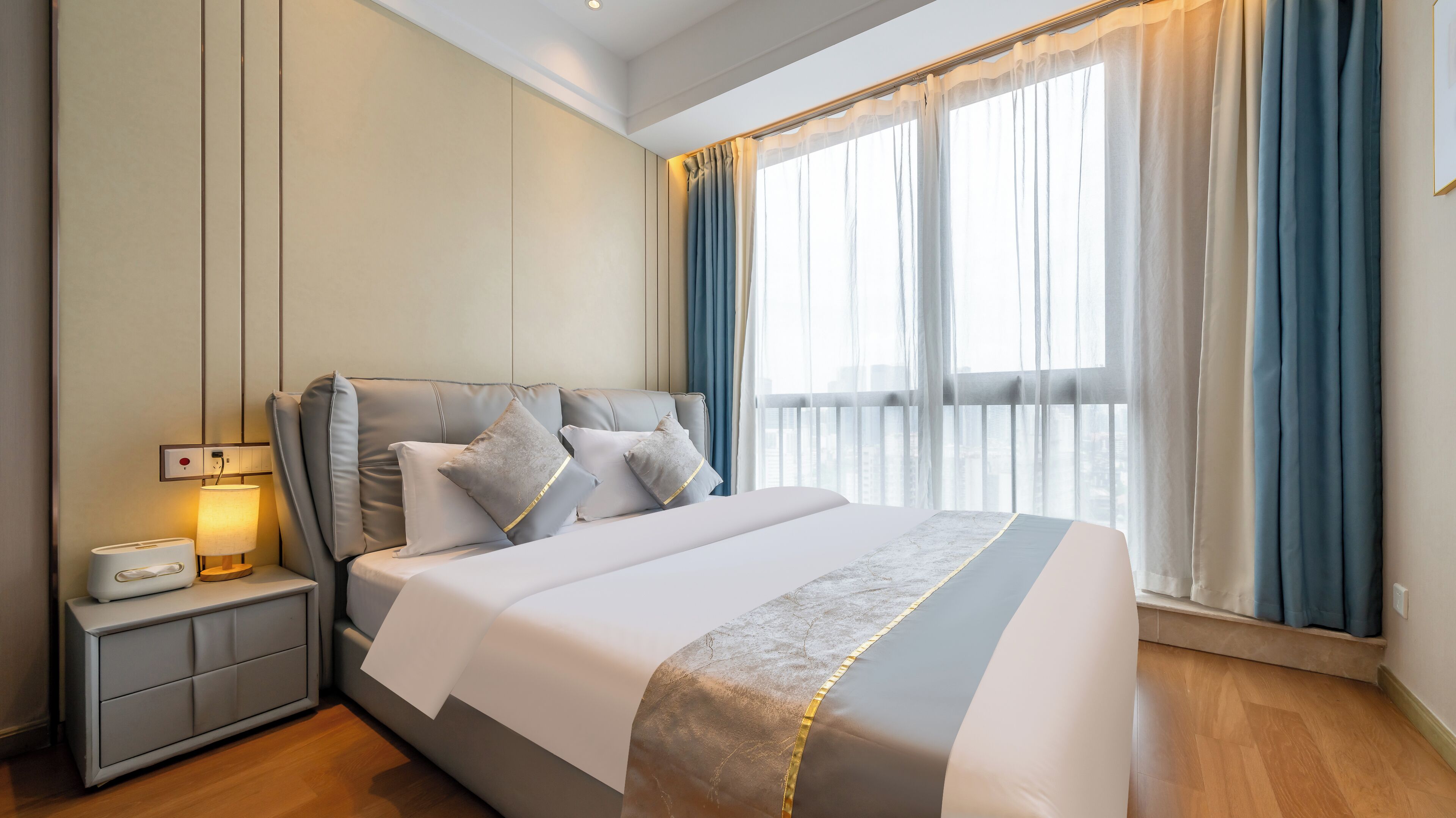 A modern hotel room with a large bed, a bedside table with a lamp, and a view of the city through large windows.