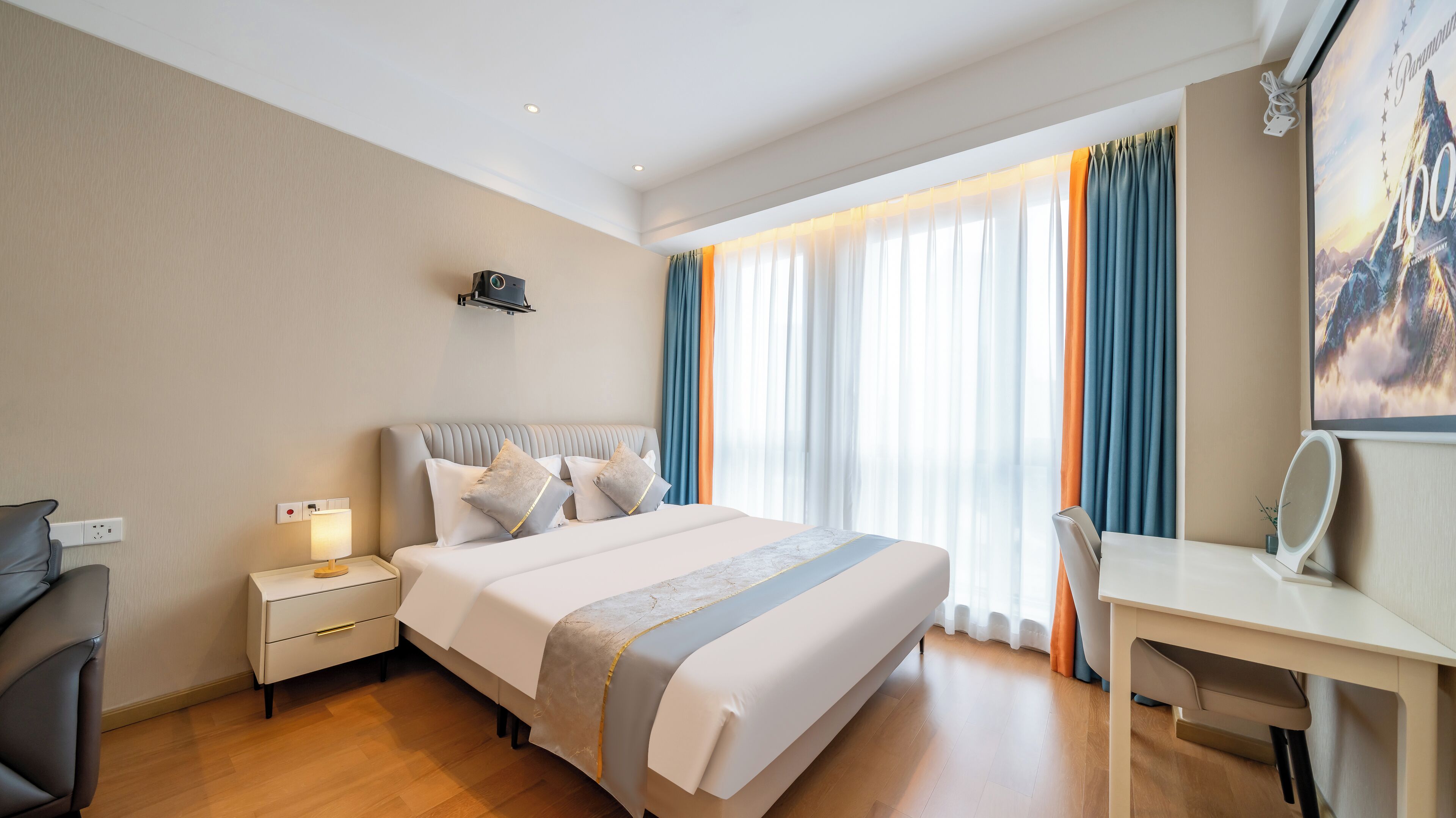 A hotel room with a bed, bedside table, lamp, desk, chair, and a large window with curtains.