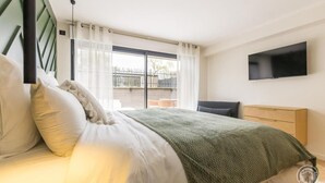 Een slaapkamer met een bed, een aan de muur bevestigde televisie en uitzicht op buiten door een groot raam.