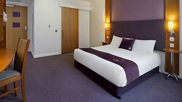 A hotel room with a large bed, a wooden chair, a desk, and a door. - Coventry South (A45) (Coventry)