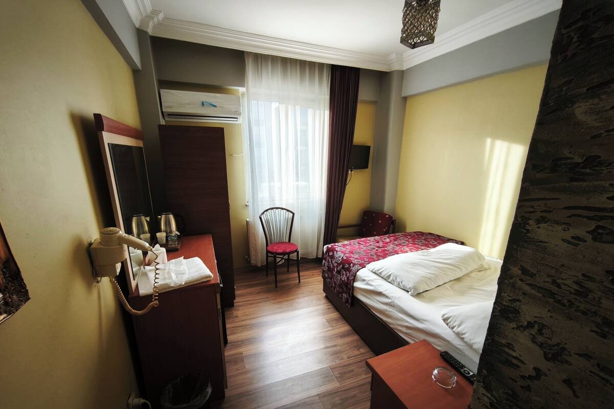 Habitación de hotel con una cama, un escritorio con teléfono, una silla y una mesita con un vaso.