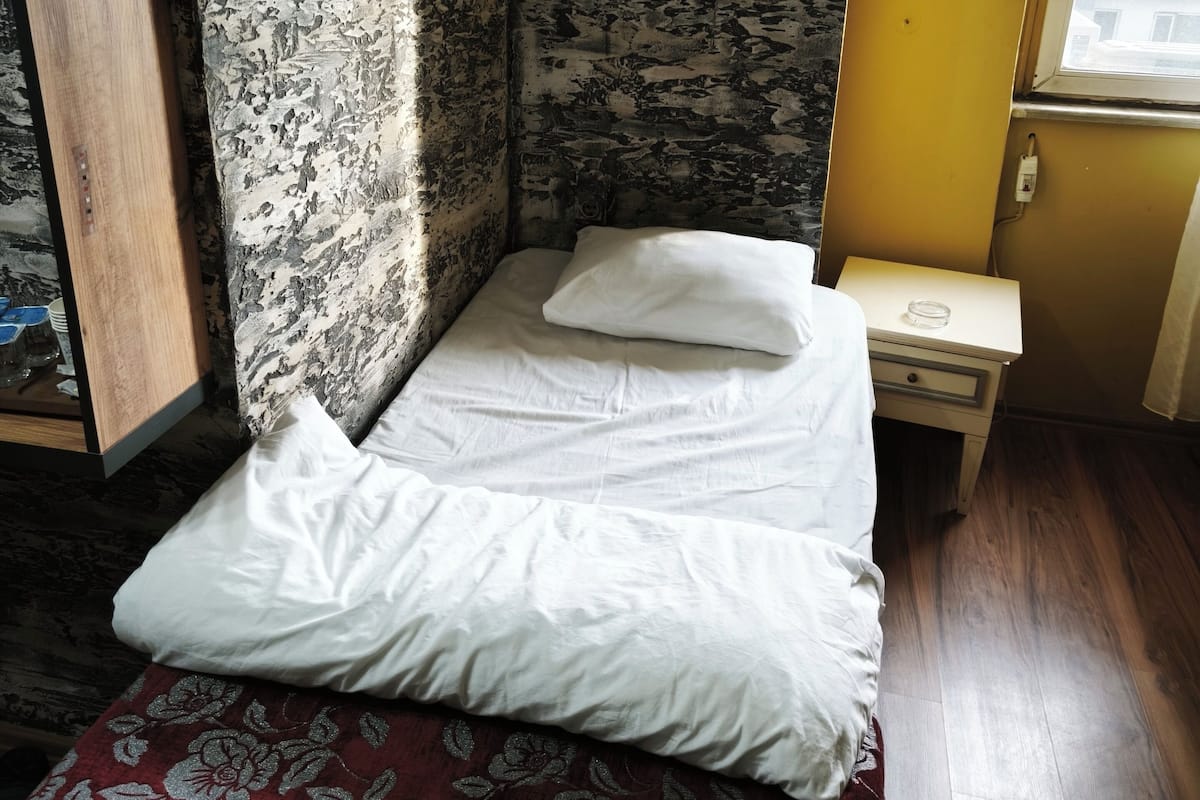 Una habitación pequeña con cama individual, pared con diseño, piso de madera y ventana con vista a edificios.