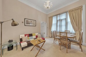 Ein Wohnzimmer mit Sofa, Couchtisch, Esstisch mit Stühlen, einer Kandelaber und einem großen Fenster mit Vorhängen.