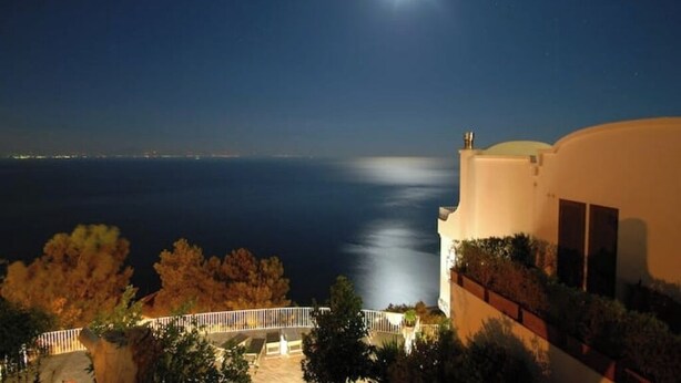 Une vue sur la côte de nuit, avec une mer illuminée par la lune, un bâtiment blanc doté d’un balcon et d’une balustrade.