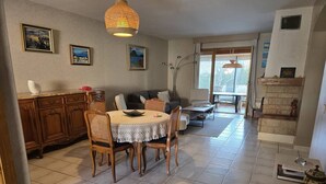 Un comedor con una mesa redonda, sillas y un centro de mesa decorativo. Se observa un sofá y una chimenea al fondo.
