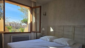 Un dormitorio con un amplio ventanal, una cama con ropa blanca, una lámpara de noche y una luminaria en la pared.