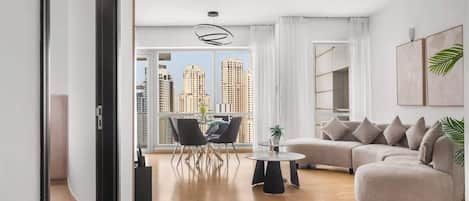 A modern living room with a curved sofa, a dining table, and chairs. There is a large window with a view of tall buildings.