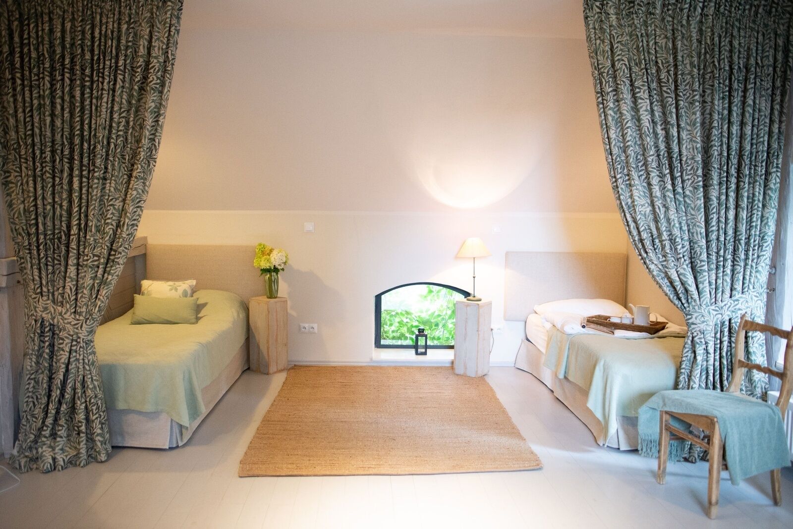 A bedroom with two beds, a window with a view, a bedside table with a lamp, and a chair.