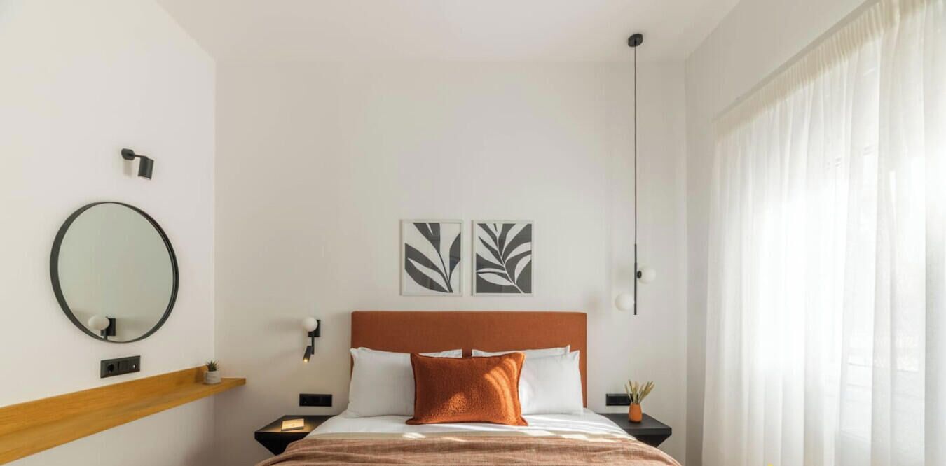 A hotel room with a bed, a round mirror, two wall-mounted light fixtures, and a window with sheer curtains.
