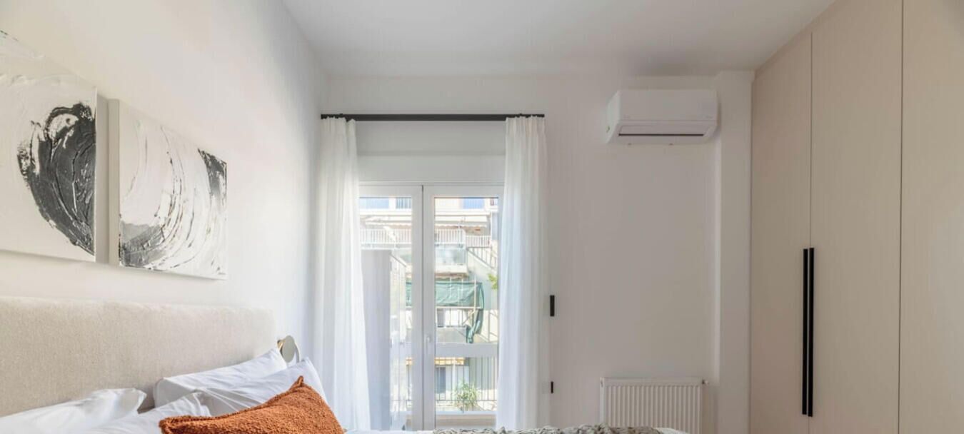 A bedroom with a bed, a window with curtains, an air conditioning unit, and a view of buildings outside.