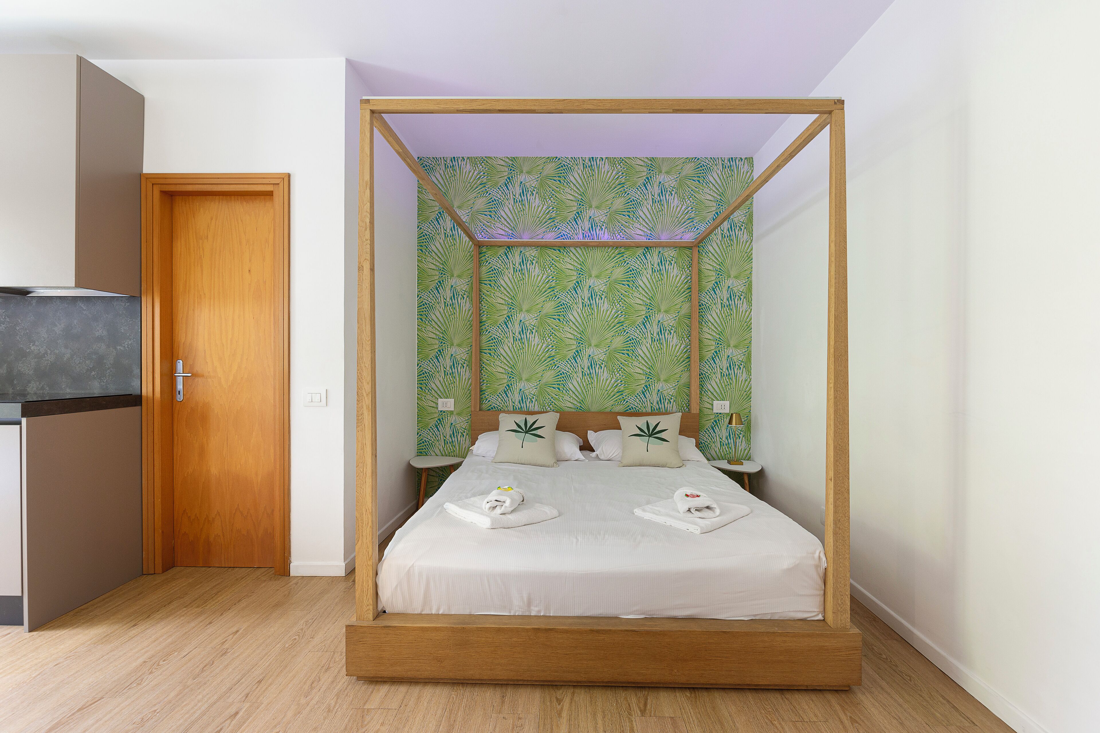 A four-poster bed with a leaf-patterned wallpaper, a wooden door, and a small bedside table.