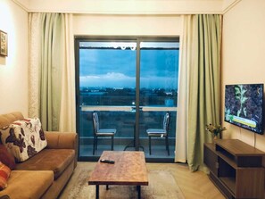 Ein Wohnzimmer mit einer Couch, einem Couchtisch und zwei Sesseln. An der Wand befindet sich ein Fernseher und ein Balkon mit Blick auf die Stadt.