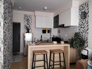 Een kleine keuken met witte kasten, een inbouwkoelkast, een houten bar met krukken, en een potplant.