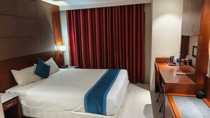 A hotel room with a bed, a desk, a chair, a nightstand, a lamp, and a large window with curtains. - BluBay Waterfront Inani (Cox's Bazar)
