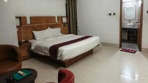 A hotel room with a bed, a nightstand, a chair, a small table, an air conditioning unit, and a bathroom visible through an open door. - BluBay Waterfront Inani (Cox's Bazar)