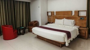 A hotel room with a large bed, a wooden headboard, a nightstand, a red chair, and a small table. - BluBay Waterfront Inani (Cox's Bazar)