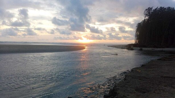 A beach scene with a sunset over the water, a sandy shore, and a line of trees in the background. - BluBay Waterfront Inani (Cox's Bazar)