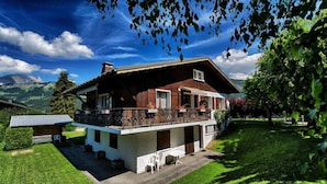 Un chalet a due piani con un balcone, immerso nel verde e con montagne sullo sfondo.