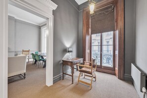 A room with a dining area, a wooden chair, a table with a lamp, and a large window with a view of buildings. - Designer Mayfair Apartment (London)