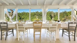 Un comedor con una mesa blanca para seis personas, sillas blancas, ventanas amplias con cortinas blancas y vistas a vegetación al exterior.
