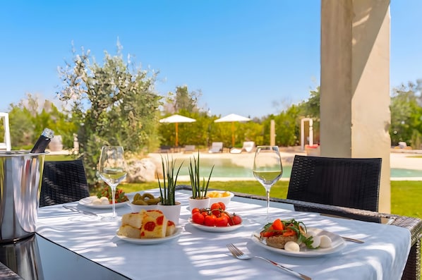 An outdoor dining table with plates of food, a wine glass, and a kettle.