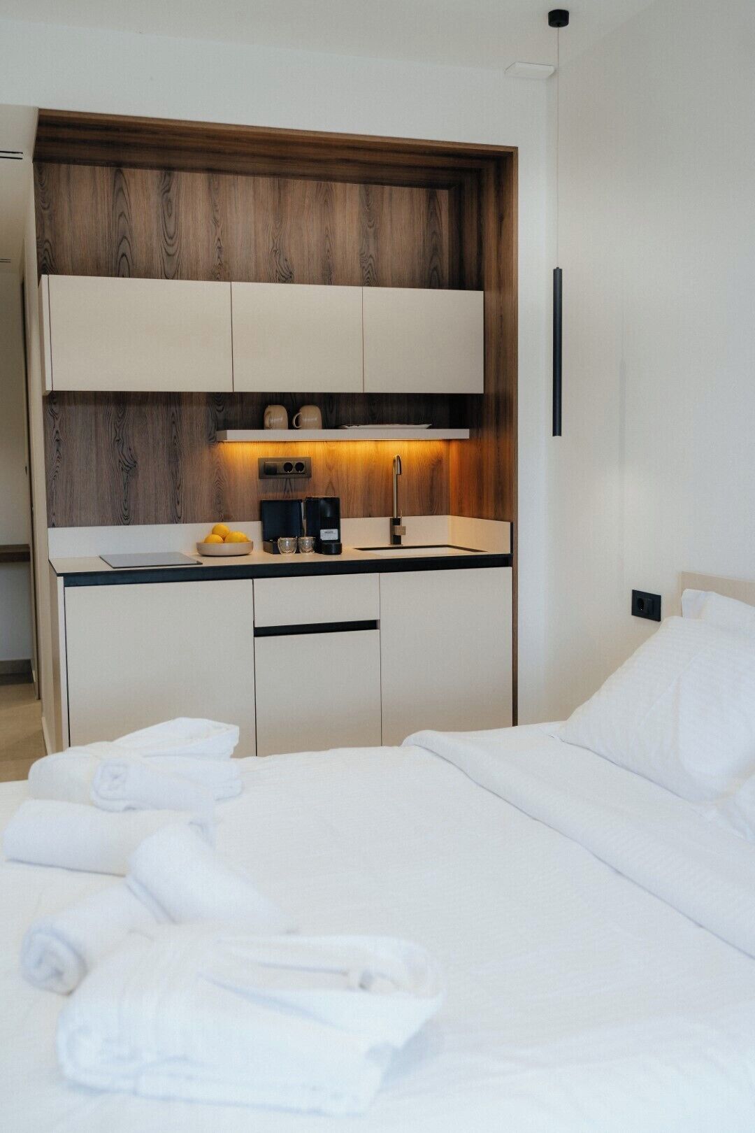 Moderni hotellihuone, jossa on siististi pedattu sänky, pieni keittokomero kahvinkeittimellä sekä yksi seinä, joka on koristeltu puupaneloinnilla.
