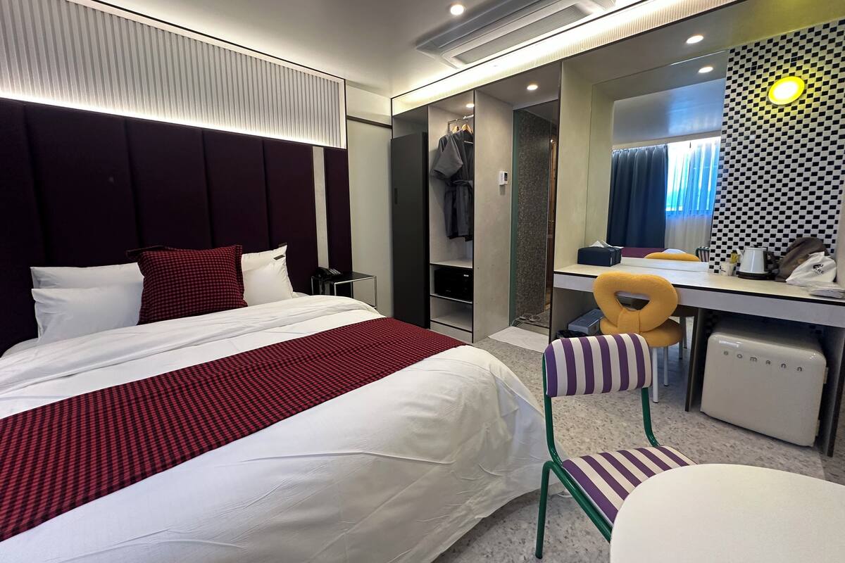 A modern hotel room with a large bed, a desk, a chair, a mirror, and a wardrobe.