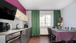 Une cuisine moderne équipée d’un téléviseur à écran plat, d’un coin repas avec une table dressée pour deux personnes, et offrant une vue sur la rue par une fenêtre.