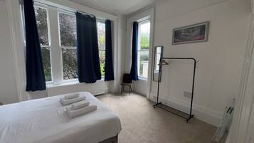 Et soveværelse med seng, radiator, stol, knagerække og et vindue med gardiner.