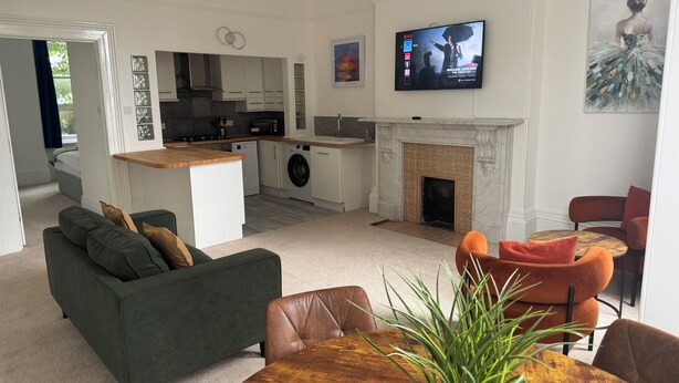 En moderne stue med sofa, spisebord, stole, et køkken med hårde hvidevarer, en pejs og et TV ophængt på væggen.