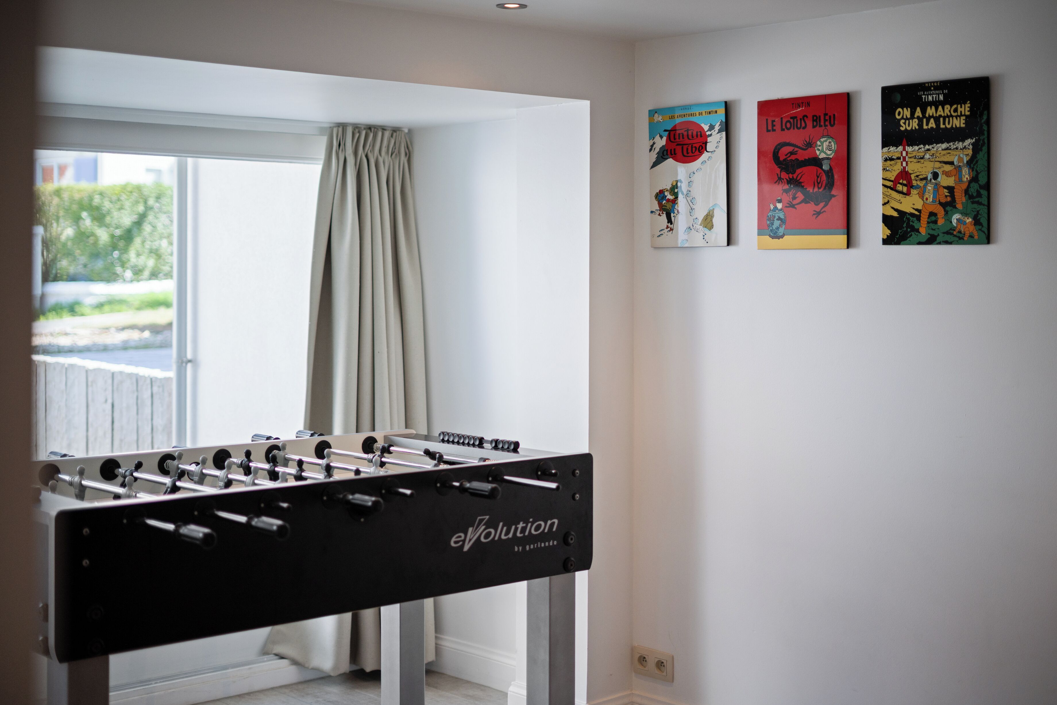 Ein Zimmer mit Kicker, einem Vorhangfenster und drei Bildern an der Wand.
