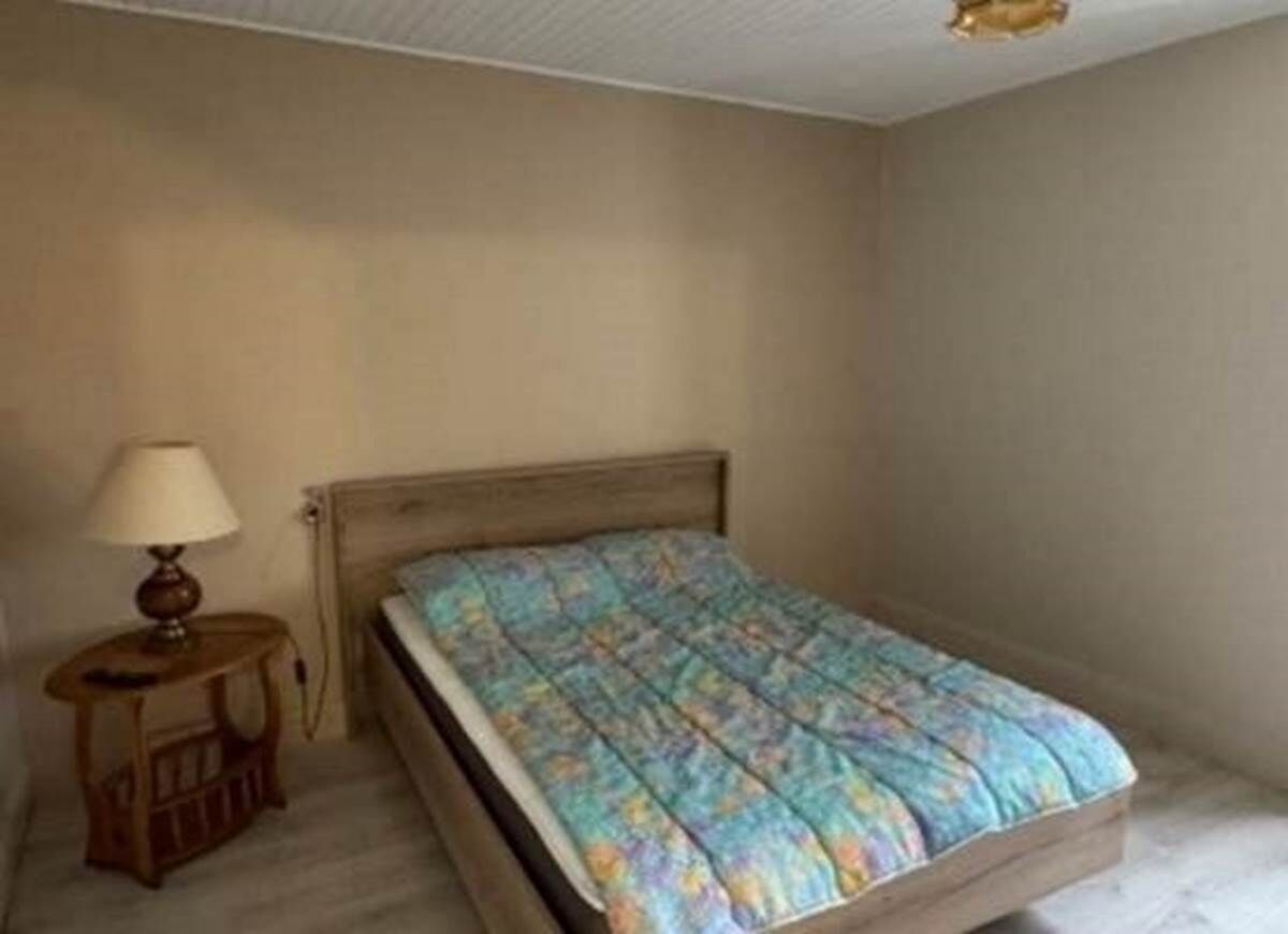 A bedroom with a wooden bed, a small bedside table with a lamp, and a plain wall.