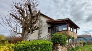 Une maison à deux étages avec une façade en pierre, un balcon en bois et un grand arbre dans la cour.